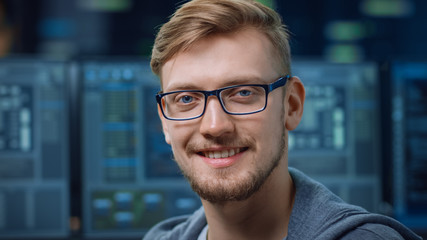 Poster - Portrait of a Smart and Handsome IT Specialist Wearing Glasses Smiles at the Camera. In the Background Unfocused Personal Computers with Screens in Technological Research Data Center