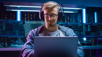 Wall Mural - Portrait of Software Developer / Hacker / Gamer Wearing Glasses and Headset Sitting at His Desk and Working / Playing on Laptop. In the Background Dark High Tech Environment with Multiple Displays.