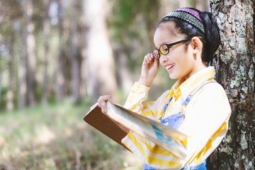 Wall Mural - Cute little girl with glasses reading book in garden