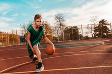 Sports And Basketball. A Young Teenager In A Green Tracksuit Plays Basketball, Leads The Ball Before Throwing. Blue Sky And A Sports Field In The Background. Horizontal