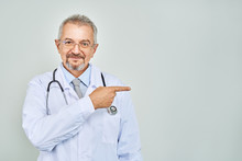 Cheerful Mature Doctor Posing And Smiling At Camera, Healthcare And Medicine. Isolate On Blue Background. Hand Pointing At Copy Space.