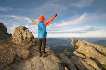 Young child boy hiker standing with raised hands in mountains enjoying view of amazing mountain landscape at sunset.