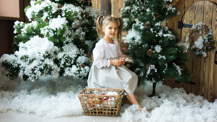 Cute little girl in a light dress sits on a stump among artificial fir trees and artificial snow with a wicker basket with toys. Children and nature.