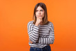 © khosrork - Portrait of worried frustrated woman with brown hair in long sleeve striped shirt standing, holding her chin and frowning, thinking with serious look. indoor studio shot isolated on orange background