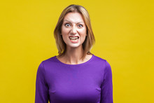 Portrait Of Beautiful Emotional Woman In Tight Purple Dress Standing With Clenched Teeth And Angry Grimace On Face, Pretending To Be Aggressive. Indoor Studio Shot Isolated On Yellow Background