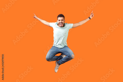 Full length of crazy overjoyed brunette man in white outfit jumping in air with raised hands, screaming loud for joy, feeling energetic and lively Poster Mural XXL