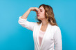 © khosrork - Awful smell! Portrait of woman with wavy hair in white jacket pinching her nose, expressing disgust to unpleasant odor, fart gases, her eyes closed with abhor. studio shot isolated on blue background