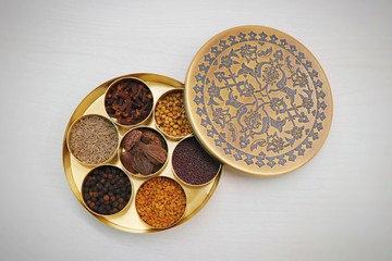 Spice box or collection of Indian spices or Garam Masala like cumin seeds, coriander, Fenugreek, big cardamom, black pepper, cloves, mustard seeds in designer brass container on wooden background