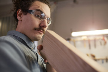 Young manual worker wearing protective eyeglasses and looking at wooden detail while making things from wood