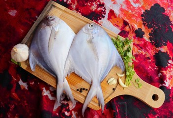 White Pomfret fish or butter fish places on a wooden board with garlic cloves, black pepper seeds and coriander