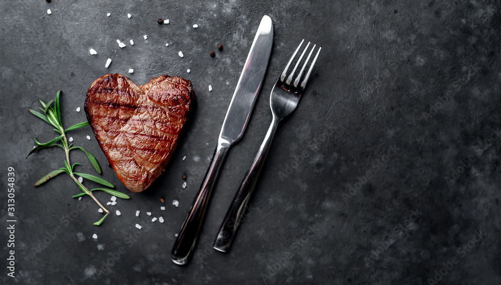 grilled beef steak in the form of a heart with spices, a knife and a fork for Valentine's Day on a stone background with copy space for your text - obrazy, fototapety, plakaty 