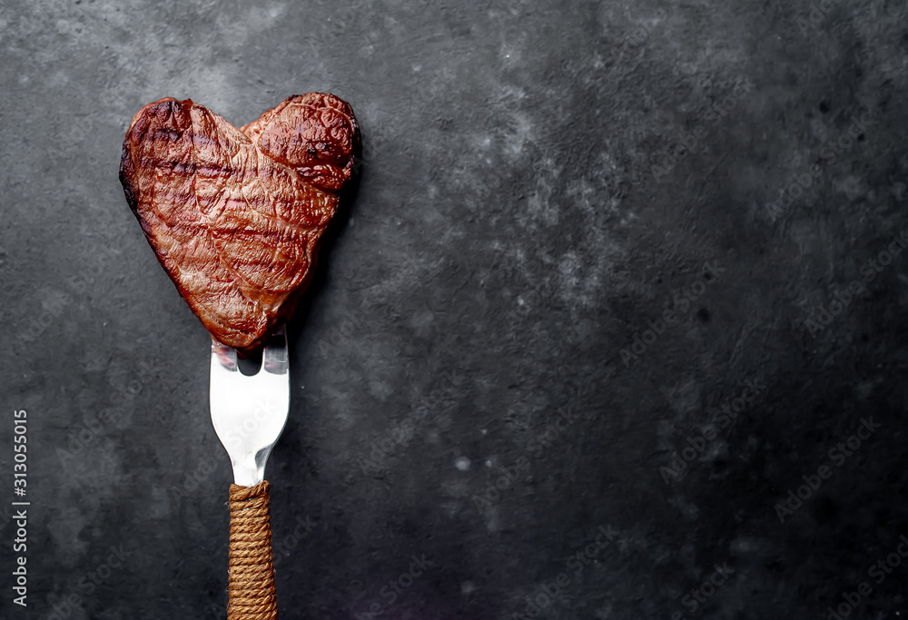 grilled beef steak in the form of a heart on a fork for Valentine's day on a stone background with copy space for your text - obrazy, fototapety, plakaty 