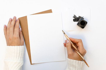 Flat lay female hand in white sweater writing wedding invitation card or love letter. Handwriting concept with an ink bottle and ink pen on a white table