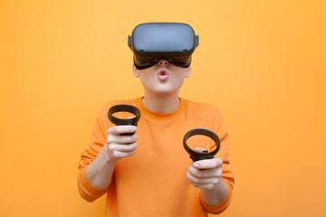 surprised guy with VR glasses on an orange background, a gamer plays a virtual game and holds virtual reality devices