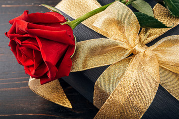 Gift wrapped in black paper and a gold ribbon. on top of the box is a luxurious red rose. The concept of an expensive gift for the holiday Valentine's Day or Women's Day March 8