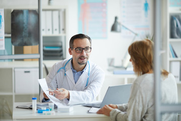 Mature healthcare worker showing some document to his patient and explaining the treatment for her while they sitting at office