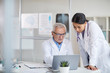© AnnaStills - Senior doctor in white coat sitting at the table working on laptop and discussing work with his nurse at office