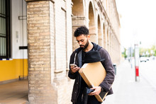 Young Man With Hat And Parcel Looking At Cell Phone In The City
