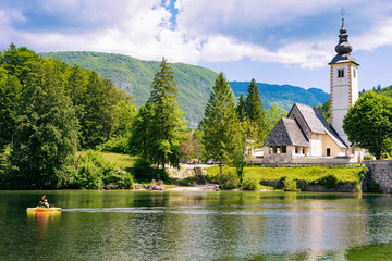 Scenery of man canoeing on Bohinj Lake, Slovenia. Nature and people kayaking, Slovenija. View of green forest, blue water. Landscape, Alpine Julian Alps mountains. John baptist church on background