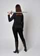 © faestock - full length portrait of a pretty brunette girl wearing a black shirt and leather boots, holding a sword. Standing pose facing away and holding a sword,  on a grey studio background.