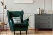 Emerald green wing back chair with pillow in grey living room interior with wooden commode