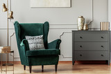 Fototapeta  - Emerald green wing back chair with pillow in grey living room interior with wooden commode