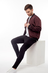 Wall Mural - Young european man in white sweater and black pants, red bomber jacket posing on white background. Isolated. Sitting.