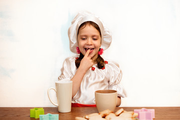 A girl in a chefs hat is standing at a kitchen table holding a cup and licks a finger