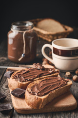 Wall Mural - Slices of bread with chocolate nut cream and a jar of cream with a cup on a wooden background. Vertical shot