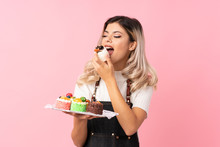 Teenager Girl Over Isolated Pink Background Holding Mini Cakes And Eating It
