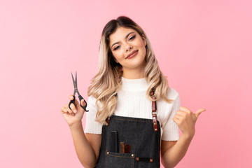 Teenager girl over isolated pink background with hairdresser or barber dress and pointing it