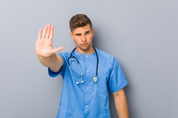 Wall Mural - Young nurse man standing with outstretched hand showing stop sign, preventing you.