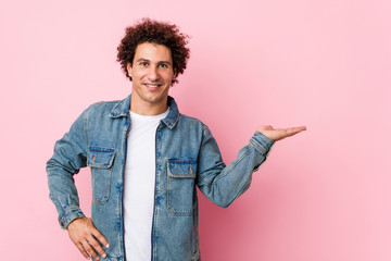 Curly mature man wearing a denim jacket against pink background showing a copy space on a palm and holding another hand on waist.