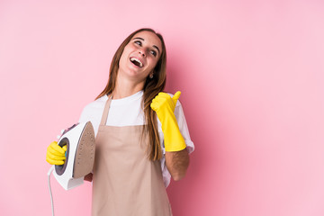 Young caucasian woman with clothes iron isolated points with thumb finger away, laughing and carefree.