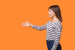 © khosrork - Side view portrait of beautiful friendly woman with brown hair in long sleeve striped shirt standing, giving hand for handshake, greeting with smile. indoor studio shot isolated on orange background