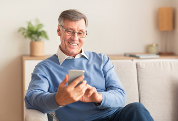 Wall Mural - Joyful Elderly Man Using Cellphone Sitting On Sofa At Home