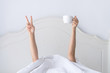 © Pattarisara - Young beautiful  woman waking up in the morning in the bed, hidding under the blanket, stretching out one's arms with a cup of coffee and showing V sign.