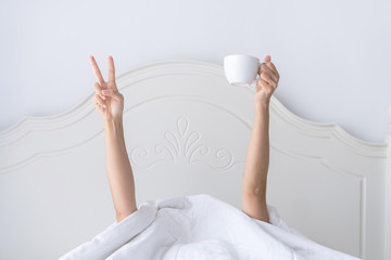 Young beautiful  woman waking up in the morning in the bed, hidding under the blanket, stretching out one's arms with a cup of coffee and showing V sign.