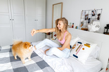 A Happy Blond Woman Is Sitting On The Bed With Her Dog Of Breed Spitz. Portrait Of A Woman And A Dog.