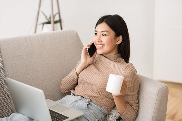 Cheerful asian girl talking on phone and drinking morning coffee