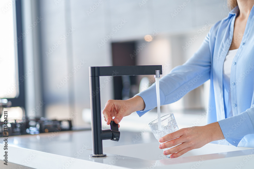 Woman pours fresh filtered purified water from a tap into a glass at kitchen at home for drinking - obrazy, fototapety, plakaty 