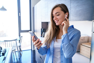 Attractive happy mobile modern woman uses wireless white headphones and smartphone for listening music and audio book at home