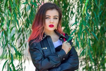 Portrait of beautiful stylish young woman with bright make-up, wearing black leather jacket, standing in a willow tree green foliage. 