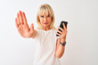 © Krakenimages.com - Middle age woman using smartphone standing over isolated white background with open hand doing stop sign with serious and confident expression, defense gesture
