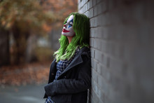 Portrait Of A Greenhaired Girl In Chekered Dress With Joker Makeup On A Brick Wall Background.