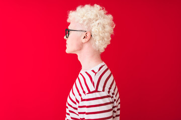 Wall Mural - Young albino blond man wearing striped t-shirt and glasses over isolated red background looking to side, relax profile pose with natural face with confident smile.