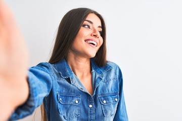 Wall Mural - Beautiful woman wearing denim shirt make selfie by camera over isolated white background looking away to side with smile on face, natural expression. Laughing confident.