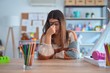 © Krakenimages.com - Young beautiful teacher woman wearing sweater and glasses sitting on desk at kindergarten tired rubbing nose and eyes feeling fatigue and headache. Stress and frustration concept.