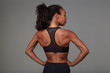 © timtimphoto - Back view of young sporty dark skinned curly woman with ponytail hairstyle holding her hands on waist and demonstrating her perfect body, standing against grey background