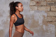 © timtimphoto - Side view of young attractive dark skinned slim woman wearing her curly long hair in ponytail hairstyle while posing over brick wall background, listening to music in her earphones